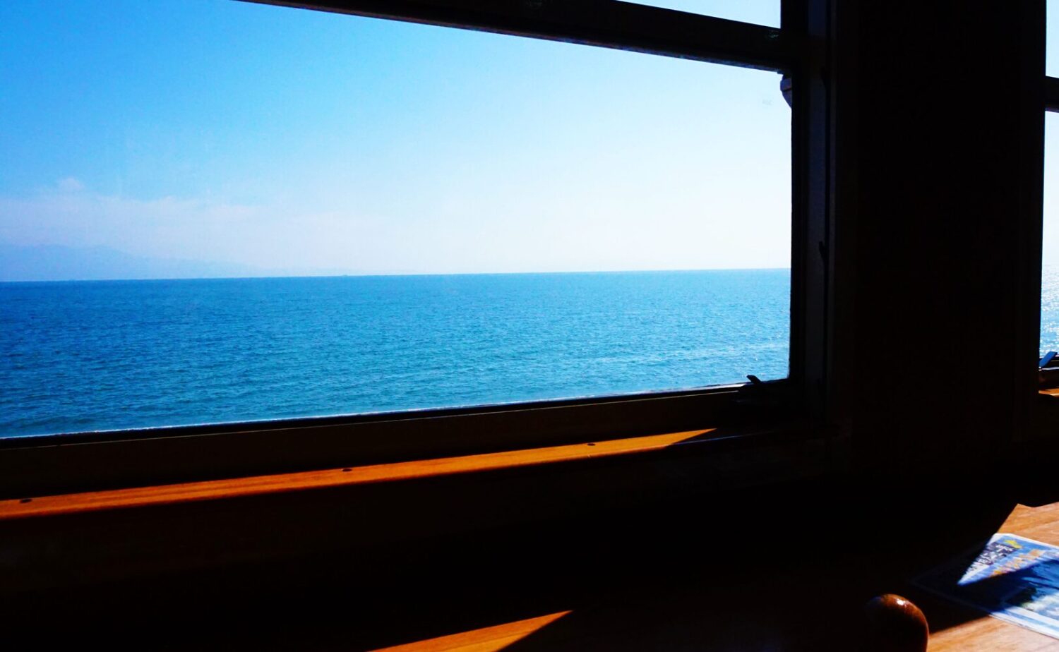 観光特急「指宿のたまて箱」のレトロな木製車窓から望む、青く澄んだ錦江湾の美しい海原と遠くに続く陸地の風景。