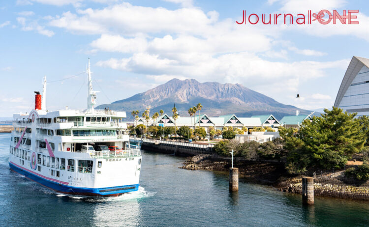 雄大な桜島を背景に鹿児島港を出港する「第二桜島丸」。白地にピンクとブルーのラインが入った船体が、青い海と空に映える美しい風景。