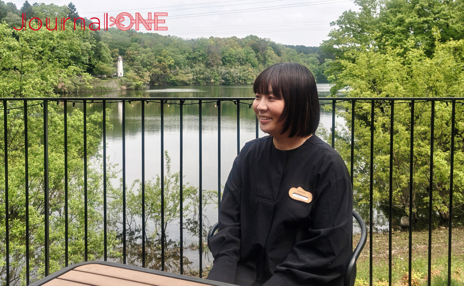 ムーミンバレーパークの宮沢湖を背景に、テラス席で微笑む「metsä」の名札をつけた羽毛田さん。背景には湖畔の豊かな緑と白い灯台が見える。