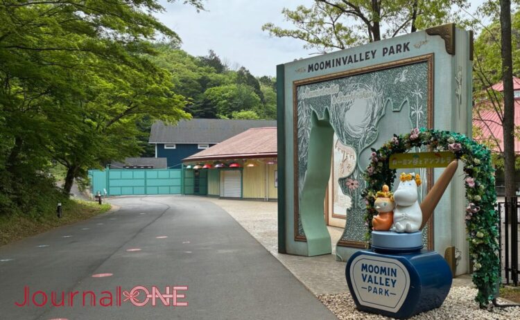 ムーミンバレーパークのエントランス。ムーミンの物語を模した巨大な本のオブジェがゲートとなり、その先には新緑の並木道が続く。「ムーミン谷とアンブレラ」の装飾を施したムーミンとリトルミイのフォトスポットが並ぶ様子。