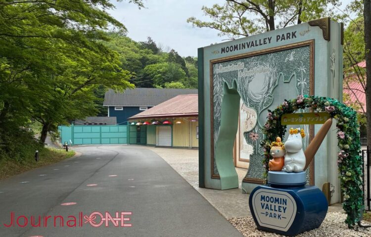 ムーミンバレーパークのエントランス。ムーミンの物語を模した巨大な本のオブジェがゲートとなり、その先には新緑の並木道が続く。「ムーミン谷とアンブレラ」の装飾を施したムーミンとリトルミイのフォトスポットが並ぶ様子。
