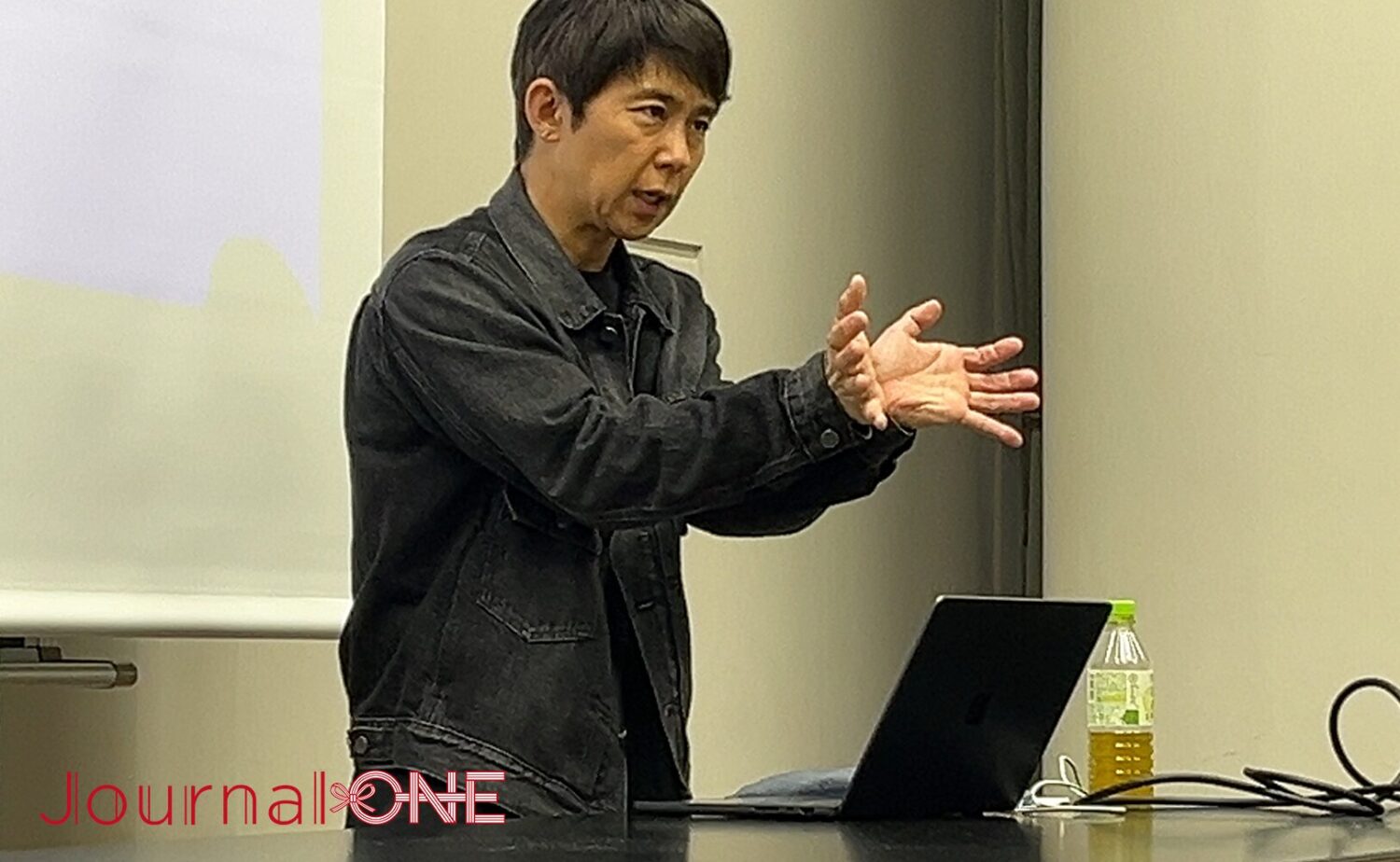 Journal-ONEの取材にて、プレゼンテーションを行う中村氏。ブラックデニムジャケットを着用し、目の前のノートPCとプロジェクタスクリーンを背に、両手を広げて概念を説明する情熱的なジェスチャーを見せている。その真剣な眼差しからは、チームラボの「不完全さ」に対する理論的かつ情緒的なアプローチと、洗練された知性が感じられる。