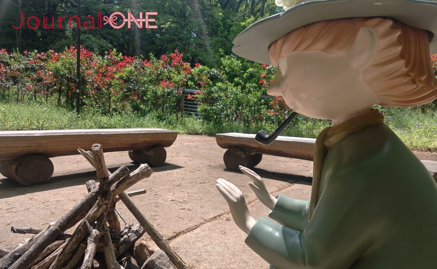 ムーミンバレーパークの「おさびし山」エリア。緑豊かな木々を背景に、パイプをくわえて焚き火にあたるスナフキンの像。横には丸太のベンチが置かれている。