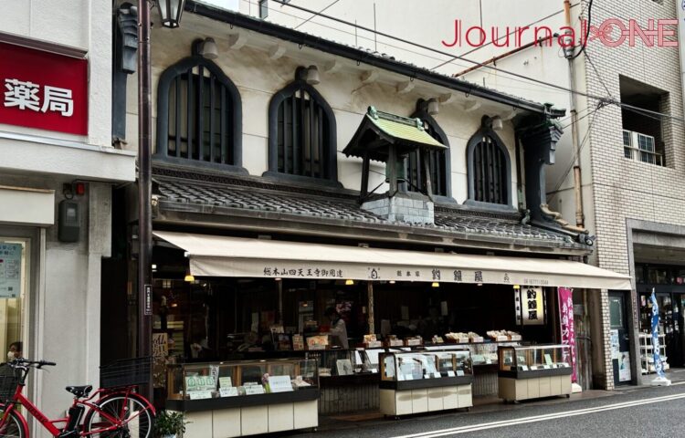 四天王寺の参道沿いに建つ和菓子店「総本家 釣鐘屋」を斜めから捉えた写真。特徴的な釣鐘型の窓と屋根の装飾が続き、手前には赤い自転車が停まっている。