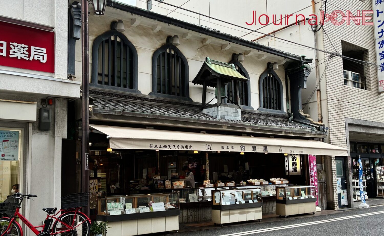 四天王寺の参道沿いに建つ和菓子店「総本家 釣鐘屋」を斜めから捉えた写真。特徴的な釣鐘型の窓と屋根の装飾が続き、手前には赤い自転車が停まっている。