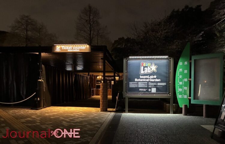長居植物園の深い夜の静寂に浮かび上がる、チームラボ ボタニカルガーデン 大阪の入場ゲート。漆黒の闇に包まれたチケットカウンターへの導線と、それを照らす「TeamLab ★ Botanical Garden」の鮮明な光のロゴ看板。人工的な光と周囲の自然(木々)が交差する、知性的な夜の入口。