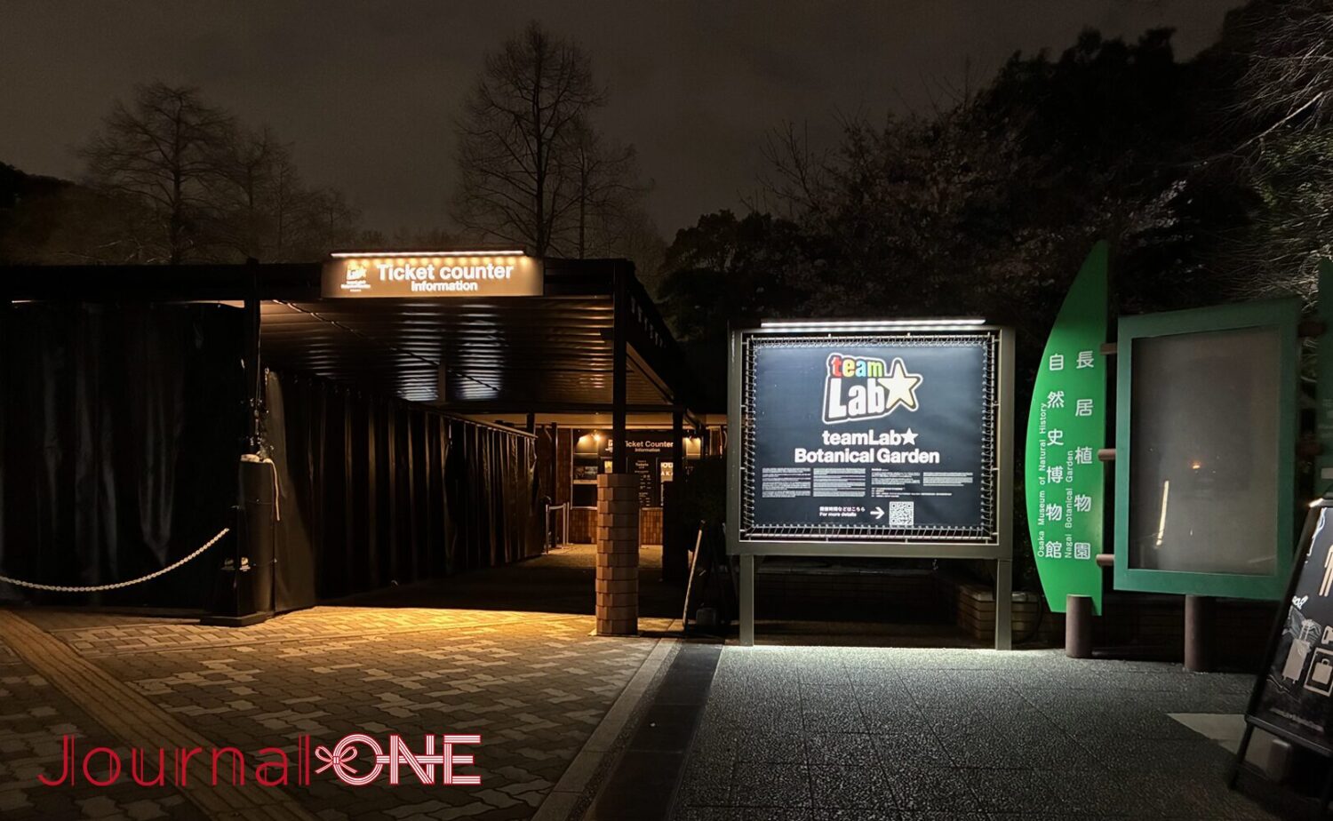 長居植物園の深い夜の静寂に浮かび上がる、チームラボ ボタニカルガーデン 大阪の入場ゲート。漆黒の闇に包まれたチケットカウンターへの導線と、それを照らす「TeamLab ★ Botanical Garden」の鮮明な光のロゴ看板。人工的な光と周囲の自然（木々）が交差する、知性的な夜の入口。