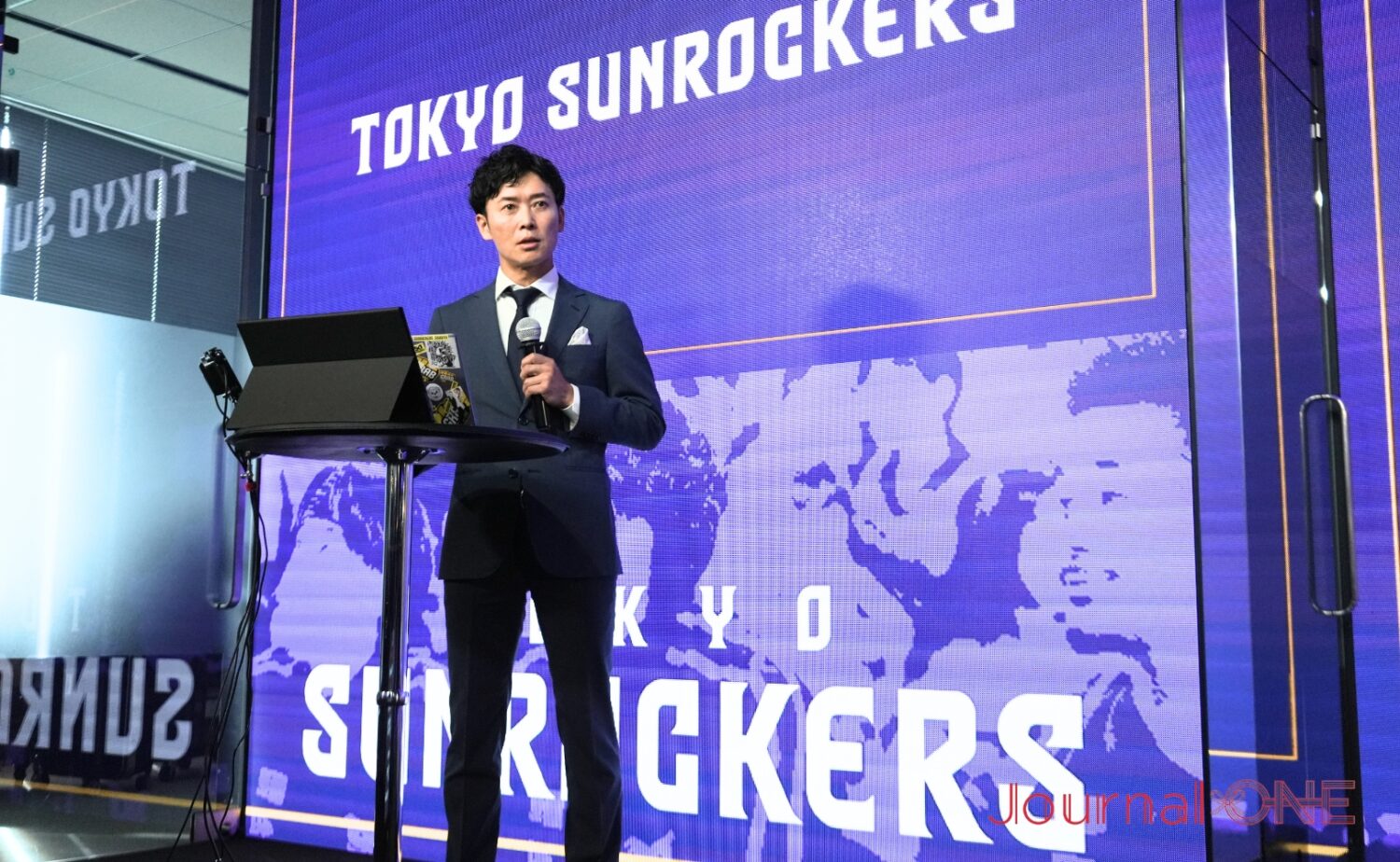 東京サンロッカーズの”紫”を背に説明する神田社長-Journal-ONE撮影